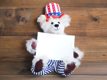 Smiling American teddy bear holding white card for 4th of July holiday on wooden backgroundの写真素材