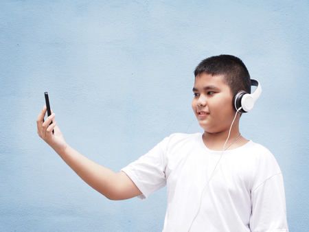asian boy with white shirt wearing headphone and playing mobile phone over blue wall background.の写真素材