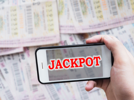 Online gambling concept.Human hand holding mobile phone with red word Jackpot on screen display over blurry Thai lottery ticket background.の写真素材
