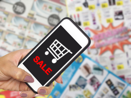 online shopping and e-commerce on internet concept. Hand holding mobile phone with cart symbol on screen display over goods catalogs brochures with promotion blurred background.の写真素材