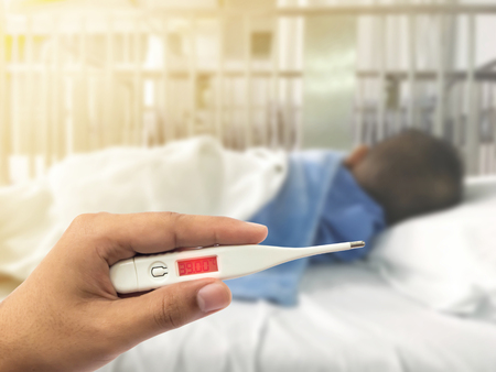 Hand holding digital thermometer with high fever over blurry illness asian kid with blue patient...