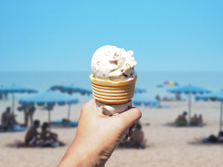 Asian woman hand holding chocolate chips ice cream cone over blurred Phuket island beach background with people sunbath on hot summer day.の写真素材