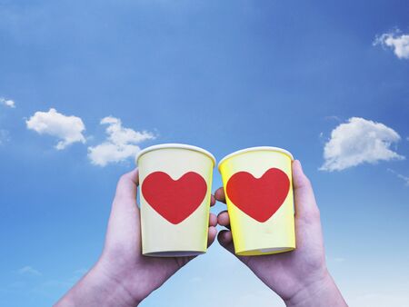 Two hands holding yellow paper take away cups of coffee with red heart shape over blue sky and copy...