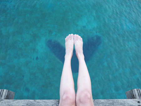 Close up beauty woman leg and bare feet sitting on wooden floor over blue sea background for...