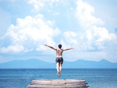 Back view of asian man jumping off wooden bridge into blue sea background. Flying freely to the sky. Summer vacation lifestyle.の写真素材