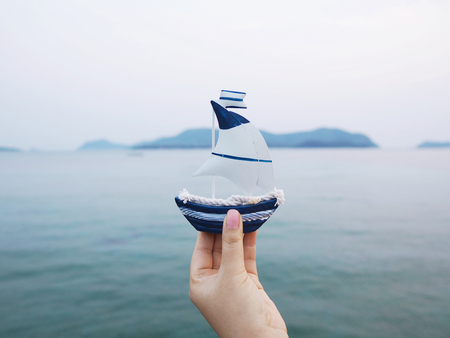Woman hand holding white sailboat model over blue sea background. Happy vacation holidays on summer beach concept.の写真素材