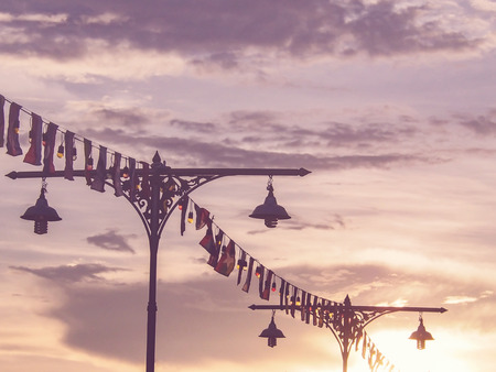 Classic Thai lamp posts with colorful light bulbs and flags for festival over dramatic sky at sunset, vintage filter effect.の写真素材