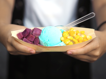 Hand holding blue homemade ice cream with purple sweet potato and corn in dried leaf cup boat shape. Healthy and save environment street food.の写真素材