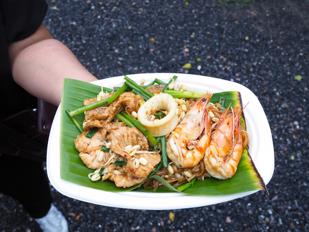 Hand holding popular street food Pad Thai for tourist. Noodles stired with seafood and pork on banana leaf over white recycle plate.の写真素材