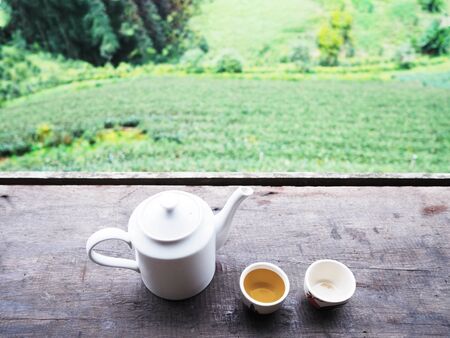 Top view of white teapot and cups on vintage wooden table over green tea farm background.の写真素材