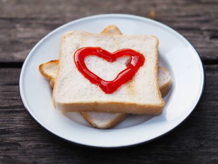 Close up toasted bread with red fruit jam heart shape on white plate over wooden background. Diet...