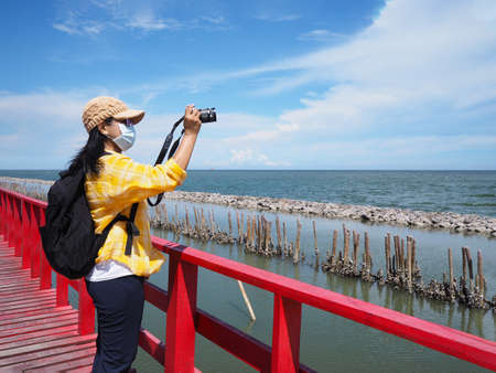 Asian woman tourist wearing face mask standing on red bridge and taking photograph summer beach at viewpoint, Samutsakhon province, Thailand. New nomal health care lifestyle concept.の写真素材