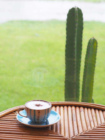 Close up cappuccino coffee cup with milk foam surface on wooden table near by glass window with green garden view.の写真素材
