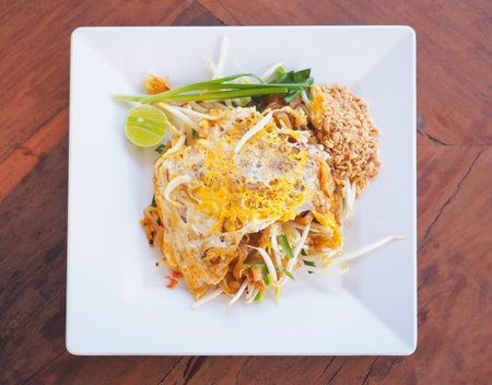 Popular Thai street food named Pad thai on white square plate placed over wooden table. Stir fried rice noodles with vegetables and egg.の写真素材