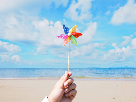 Close up hand holding pinwheel over summer beach background. Children colorful wind turbine toy.