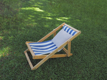 Wooden deckchair with white pillow on green grass backyard under big tree shade.