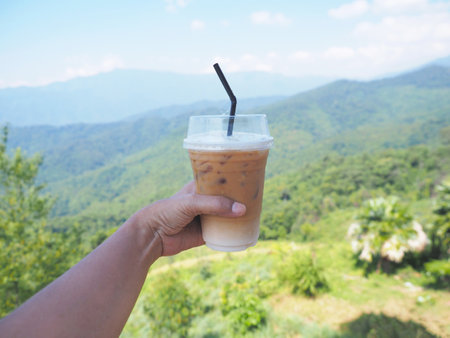 Hand holding take away glass of iced coffee over green mountain blurred background.の写真素材