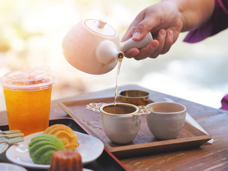 Hand holding teapot pouring hot tea through stainer into a cup and cupcake on a wooden table at Japanese cafe. Afternoon tea break.の写真素材