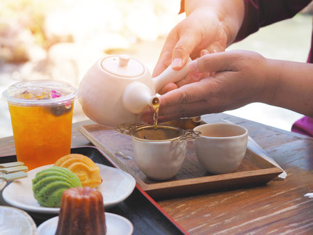 Hand holding teapot pouring hot tea through stainer into a cup and cupcake on a wooden table at Japanese cafe. Afternoon tea break.の写真素材