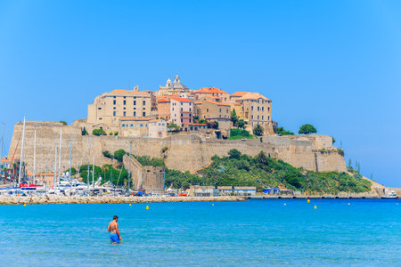 CALVI, CORSICA ISLAND - JUN 28, 2015: man relaxing in sea water and view of citadel with houses in Calvi port in backgroundのeditorial素材
