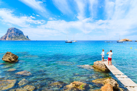 IBIZA ISLAND, SPAIN - MAY 18, 2017:  man taking picture of woman standing on small wooden pier in Cala d'Hort bay and view of Es Vedra island, Ibiza island, Spain.のeditorial素材