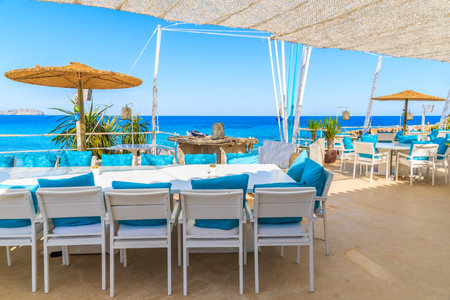 CALA NOVA BEACH, IBIZA ISLAND - MAY 20, 2017: White table and chairs with blue pillows on restaurant terrace on Cala Nova beach with beautiful sea view, Ibiza island, Spain.のeditorial素材