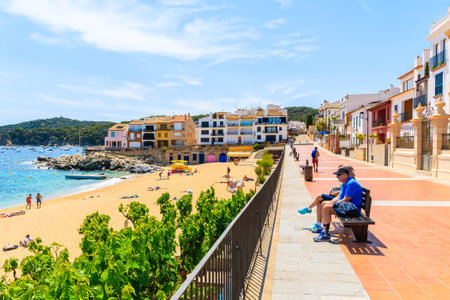 CALELLA DE PALAFRUGELL, SPAIN - JUN 6, 2019: Couple of tourists sitting on beach on coastal promenade near Canadell beach in Calella de Palafrugell village, Costa Brava, Catalonia, Spain.のeditorial素材