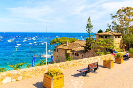 CALELLA DE PALAFRUGELL, SPAIN - JUN 6, 2019: Tourists sitting on coastal promenade in scenic fishing village with small castle and sandy beach with clear blue water, Costa Brava, Catalonia, Spainのeditorial素材