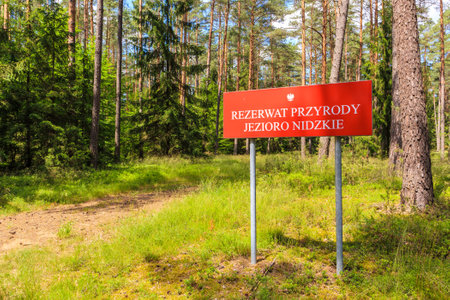 Red sign "Rezerwart Przyrody Jezioro Nidzkie" which translation is "Nature Reserve Lake Nidzkie" near forest cycling path in Puszcza Piska, Mazury Lake District, Polandのeditorial素材