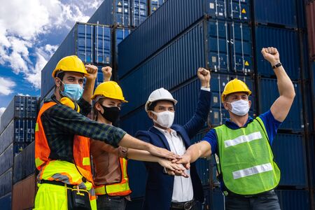Industrial workers or engineers wearing Coronavirus or COVID-19 protective masks join hands and stand in front of the shipping container.の写真素材