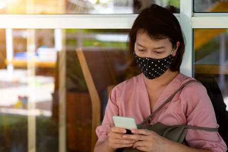 Asian woman wearing protective mask on her face while being and using smartphone in the coffee shop during virus COVID-19 pandemic.の写真素材