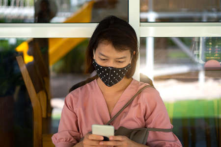 Asian woman wearing protective mask on her face while being and using smartphone in the coffee shop during virus COVID-19 pandemic.の写真素材