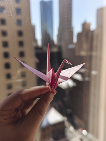 A pink origami crane, delicately held, looks optimistic against cityscape backdrop, evoking sense of peace and hopeの素材