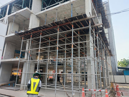 Worker in safety vest inspects steel scaffolding at concrete building under construction, urban site atmosphere with tools and caution conesの写真素材