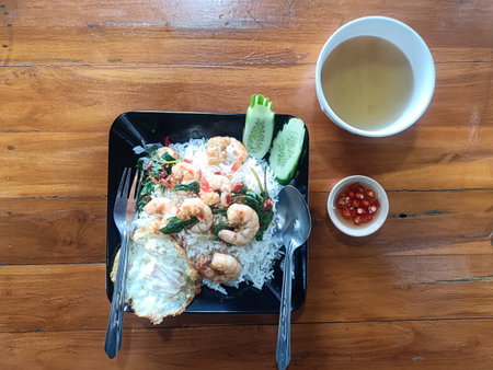 Stir fried shrimp with basil served over white rice with fried egg, cucumber slices, chili sauce and clear soup on wooden table, appetizing casual mealの写真素材