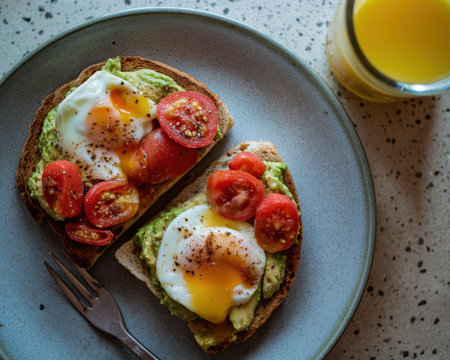 Creamy avocado toast with soft poached egg and cherry tomato slices sprinkled with pepper, served on rustic bread with orange juice nearbyの素材