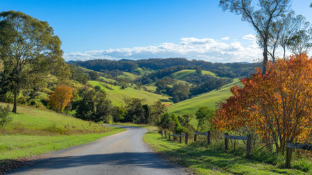 A scenic view of a picturesque road winding through lush green hills, with vibrant autumn foliage lining the sides and a clear blue sky above.の素材