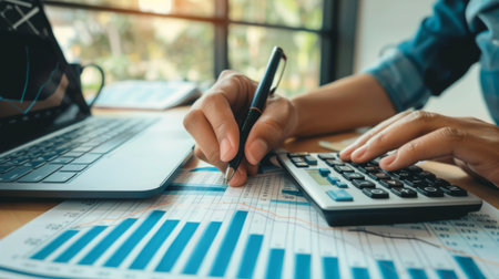 A hand writing notes on a financial report with graphs and charts, with a calculator and laptop nearby.の素材