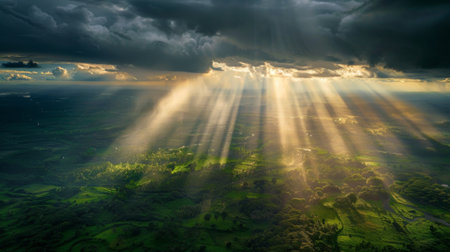 Sunlight breaking through dark rain clouds, casting a surreal glow over a lush green landscape below.の素材