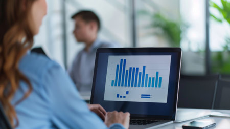 A professional explaining a bar graph to a colleague, using a laptop in a modern office setting.の素材