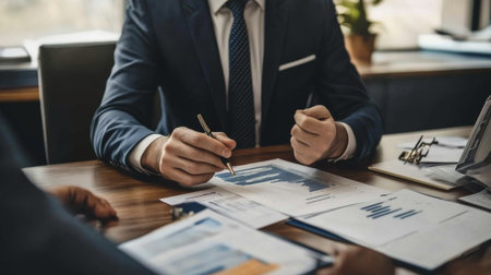 A professional banker in a suit discussing financial strategies with a client across a desk, with charts and financial reports visible in the background.の素材