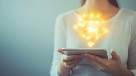 Person holding a tablet with a glowing idea icon on the screen, set against a clean, white background to highlight the focus on innovative thinking.の素材