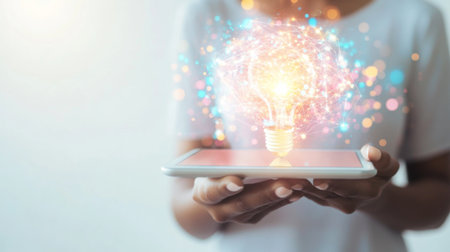 Person holding a tablet with a glowing idea icon on the screen, set against a clean, white background to highlight the focus on innovative thinking.の素材