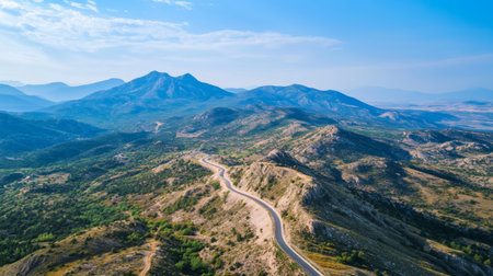 Aerial view of a mountain road snaking along a ridge, with a vast expanse of mountainous terrain and a clear blue sky in the background.の素材