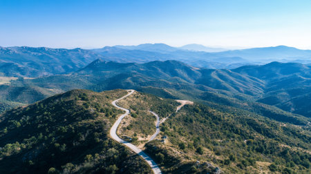 Aerial view of a mountain road snaking along a ridge, with a vast expanse of mountainous terrain and a clear blue sky in the background.の素材