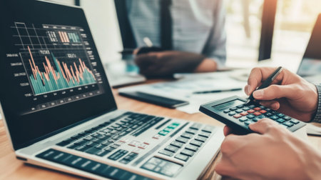 A financial advisor using a calculator to explain investment options to a client, with charts and graphs displayed on a laptop screen in the background.の素材