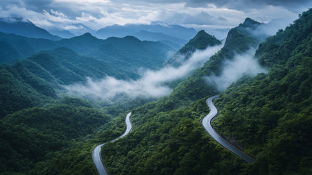 A winding road meanders through lush green mountains, enveloped in mist and clouds. A tranquil scene ideal for travel and nature enthusiasts.の素材