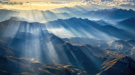 An aerial view of expansive mountain ranges with sharp peaks and deep valleys, as sunlight streams through the clouds, illuminating the rocky terrain.の素材