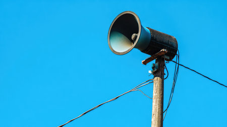 A large outdoor loudspeaker mounted on a tall pole against a clear blue sky, with cables running down, symbolizing a public announcement system.の素材