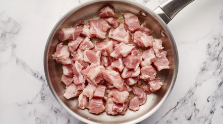 Chopped raw pork cubes lay in a stainless steel frying pan on a marble countertop, ready for cooking. Ideal for food preparation and recipe development.の素材